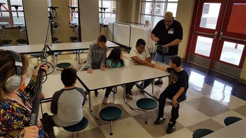 A group of children and adults are gathered around a table in a school cafeteria, engaged in a discussion while a person with a camera captures the moment.