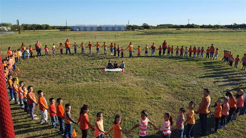 A large group of children and adults wearing orange shirts are holding hands in a circle on a grassy field, with a few individuals sitting in the center.