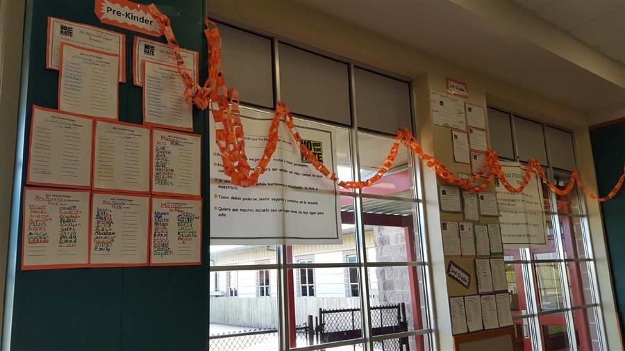 A classroom wall decorated with colorful paper chains and various student projects displayed in frames, with large windows in the background.