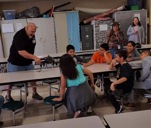 A group of children and adults are gathered in a classroom, with a man demonstrating something to the children seated at tables, while a woman observes in the background.