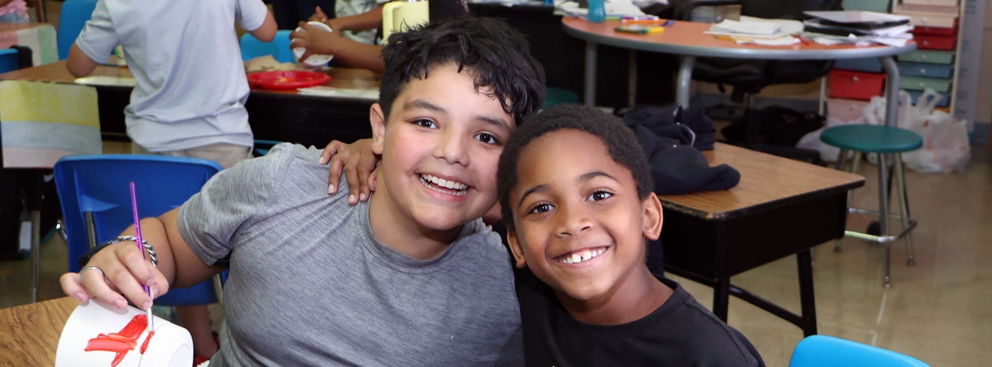 Two Hemphill students posing and smiling at the camera in a classroom with other students in the background.