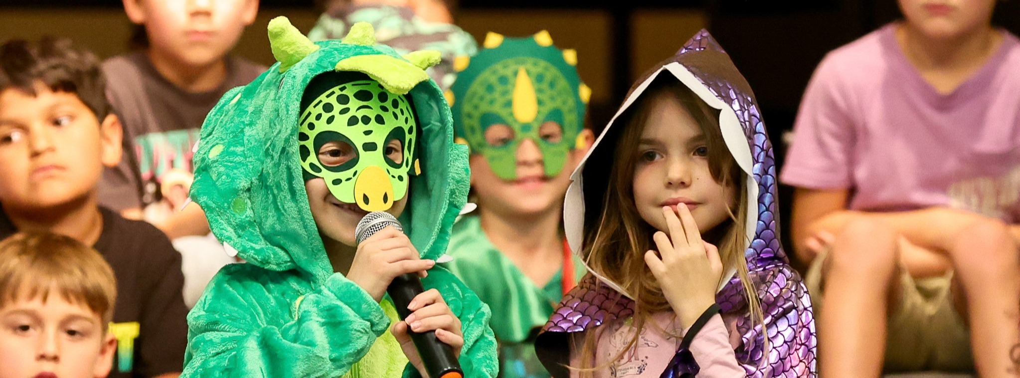 Two young students standing next to each other while wearing costumes with other students in the background.