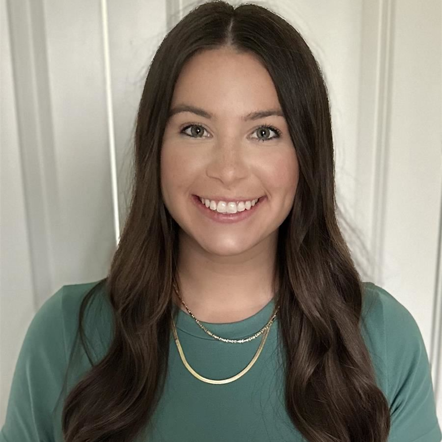 A young woman with long, wavy brown hair smiles at the camera while wearing a green shirt and layered necklaces, standing in front of a white wall.