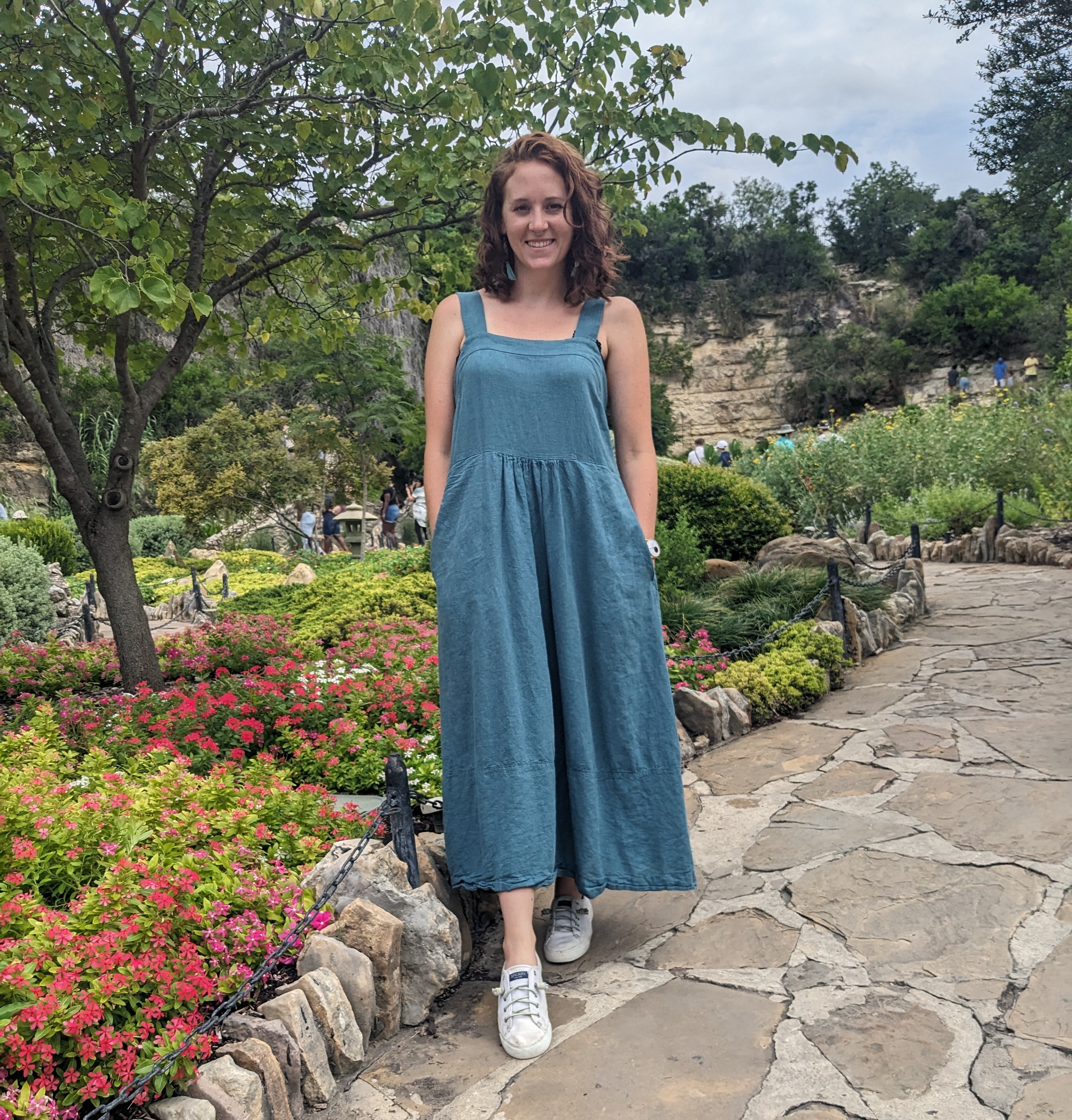 A woman with shoulder length hair in a blue dress standing along a trail in front of a bed of flowers and trees in the background.