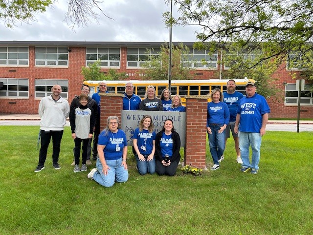 14 staff members that all attended west middle school. Each are wearing a blue WMS alumni tshirt stating the year they graduated from West.