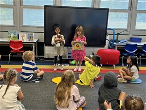 Two children stand in front of a group of seated classmates, one holding a toy and the other displaying a colorful poster, in a classroom setting.