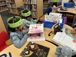 Two children wearing green hats are seated at a table in a library, engaged in reading books, with various colorful books and stuffed animals scattered around them.