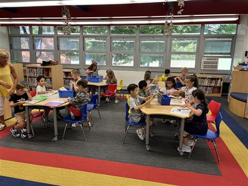 A classroom filled with children engaged in activities at tables, with large windows providing natural light and colorful rugs on the floor.