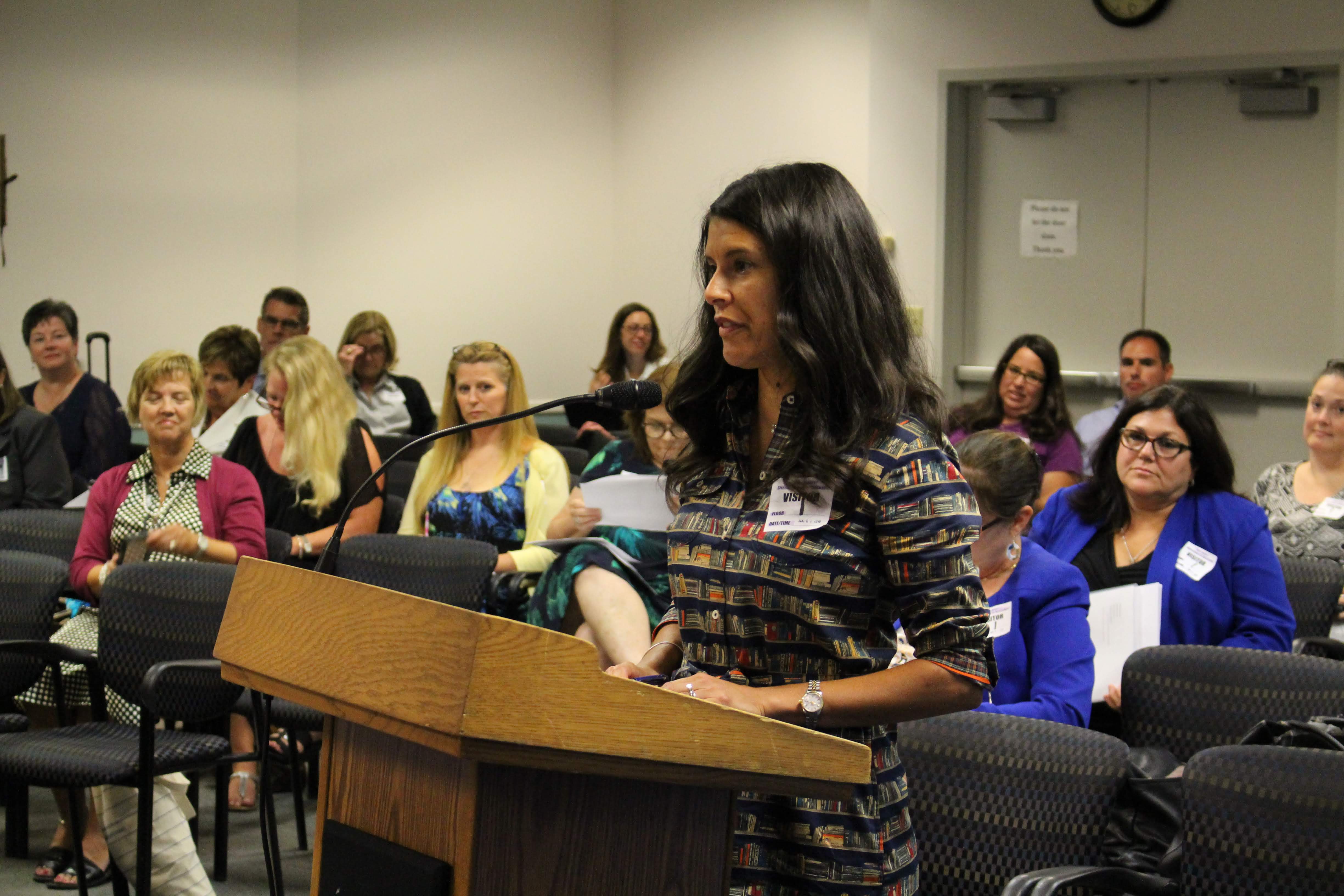 A woman stands at a podium speaking to an audience in a meeting room filled with seated attendees.