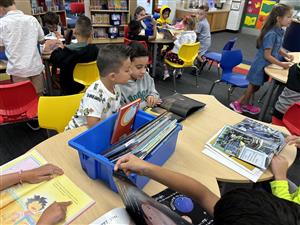 A classroom scene showing children engaged in reading and exploring books at a table, with colorful chairs and a bookshelf in the background.