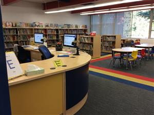 A view of a library interior featuring a curved circulation desk with computers, surrounded by bookshelves and a colorful carpet leading to a children's seating area with small tables and chairs.