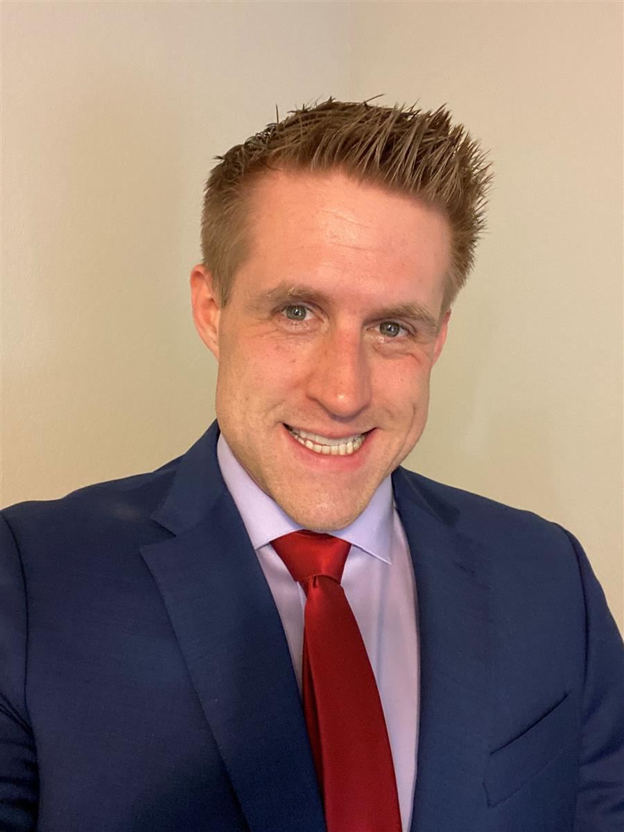 A man with short, spiky hair is smiling while wearing a dark suit, light purple shirt, and a red tie, posing against a plain background.