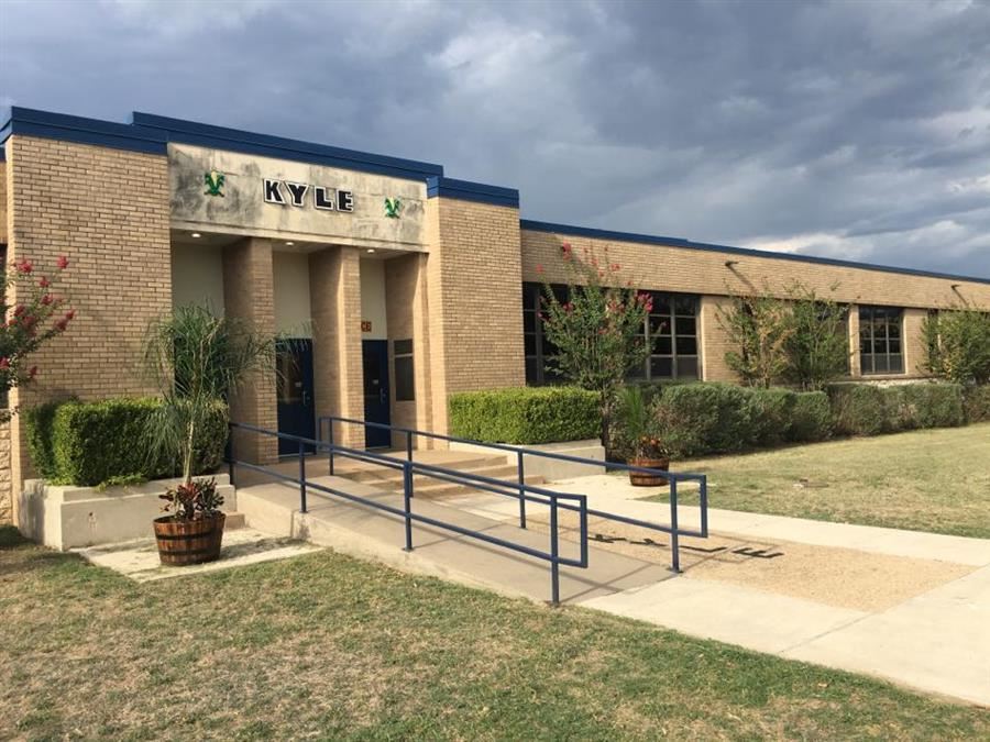 A brick building with the name 'KYLE' displayed above the entrance, featuring a ramp and landscaped areas in front, under a cloudy sky.
