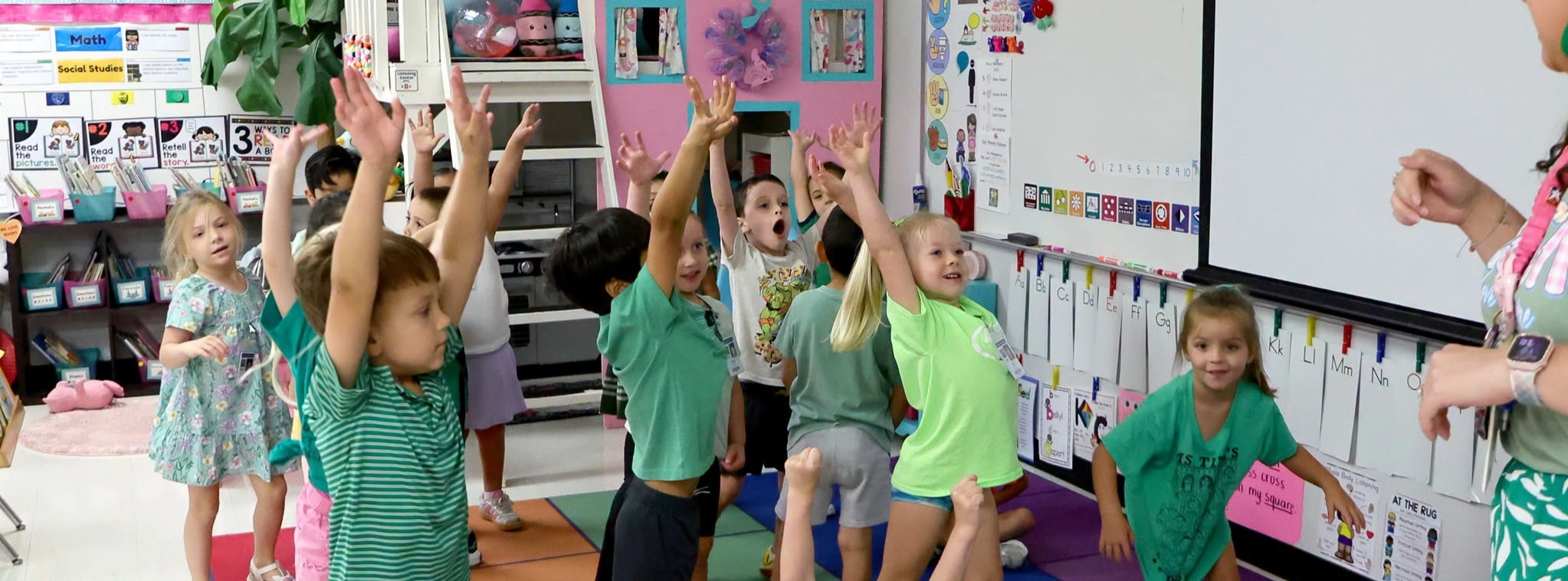 A classroom of students with the majority of students wearing green shirts and with their hands up.