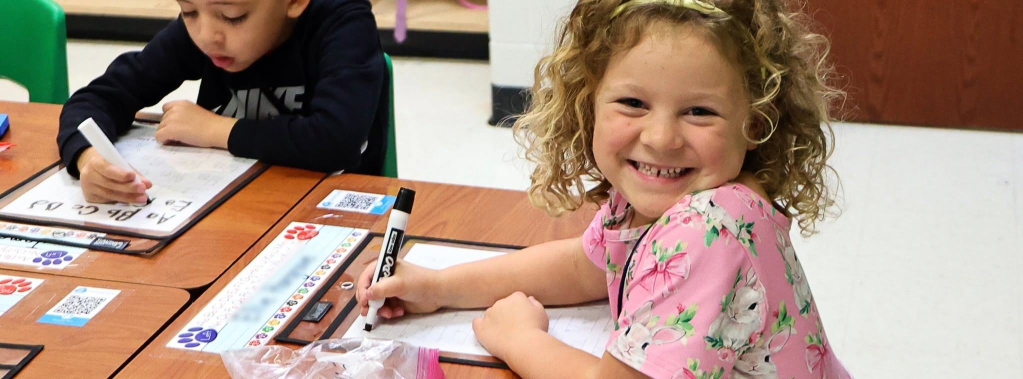 A girl with curly hair smiling at the camera while doing classwork.