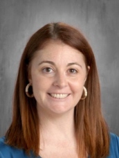 A smiling woman with shoulder length red hair, and hoop earrings wearing a blue shirt against a gray background..