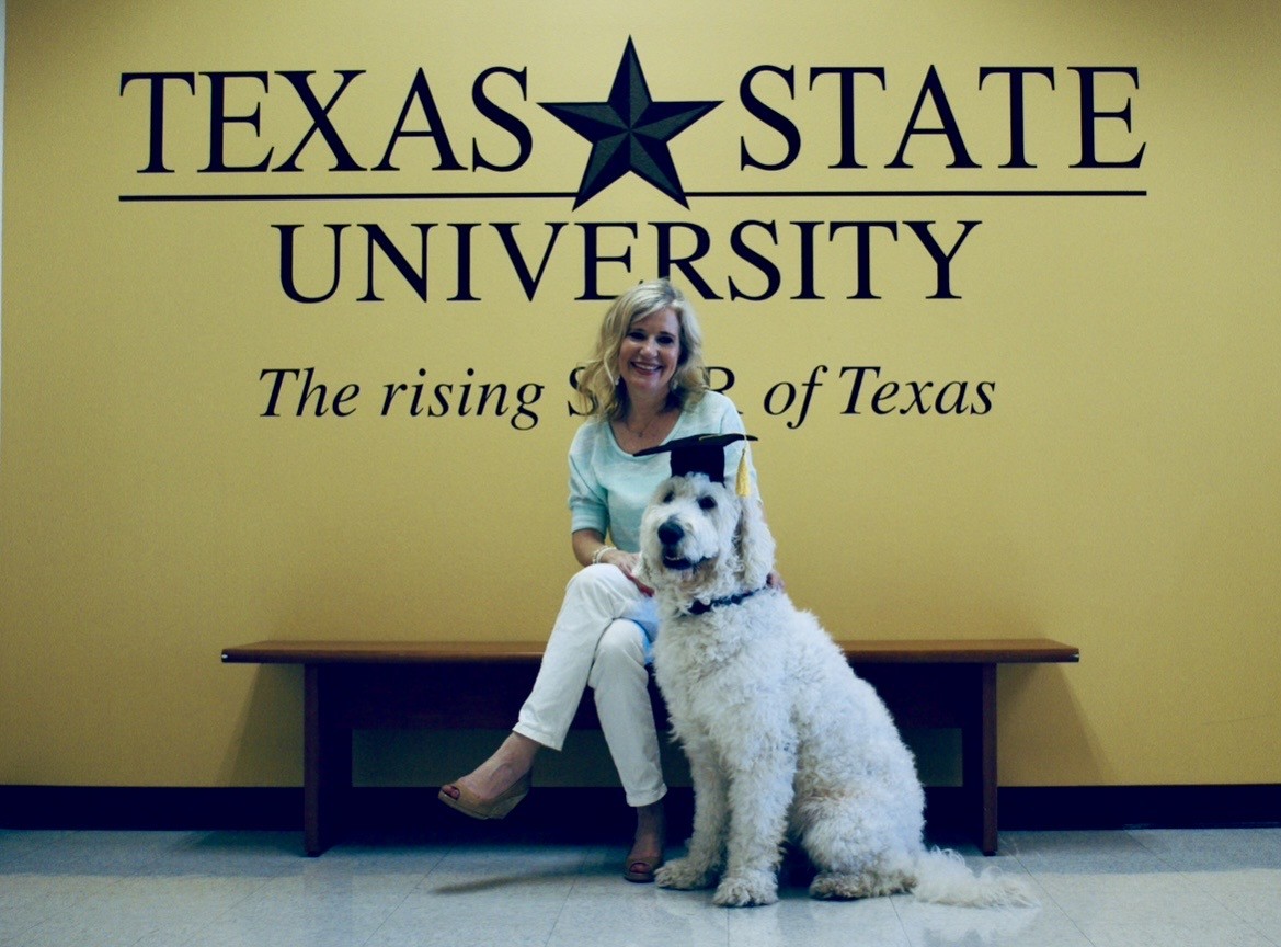 A woman sitting on a bench in front of the Texas State University logo with her dog.