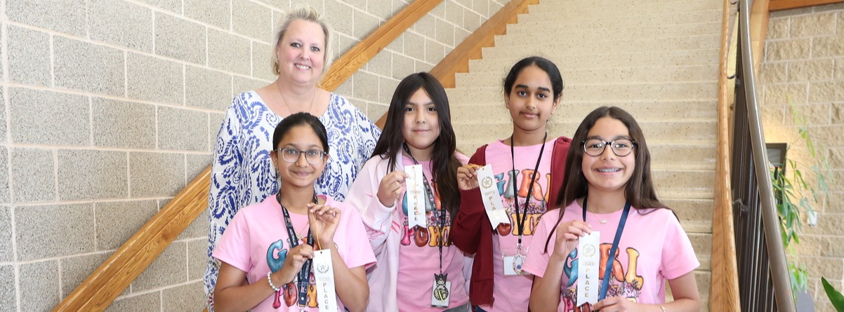a group of four students wearing matching pink shirts while showcasing their white ribbon, next to the students is a smiling woman in a blue and white shirt.