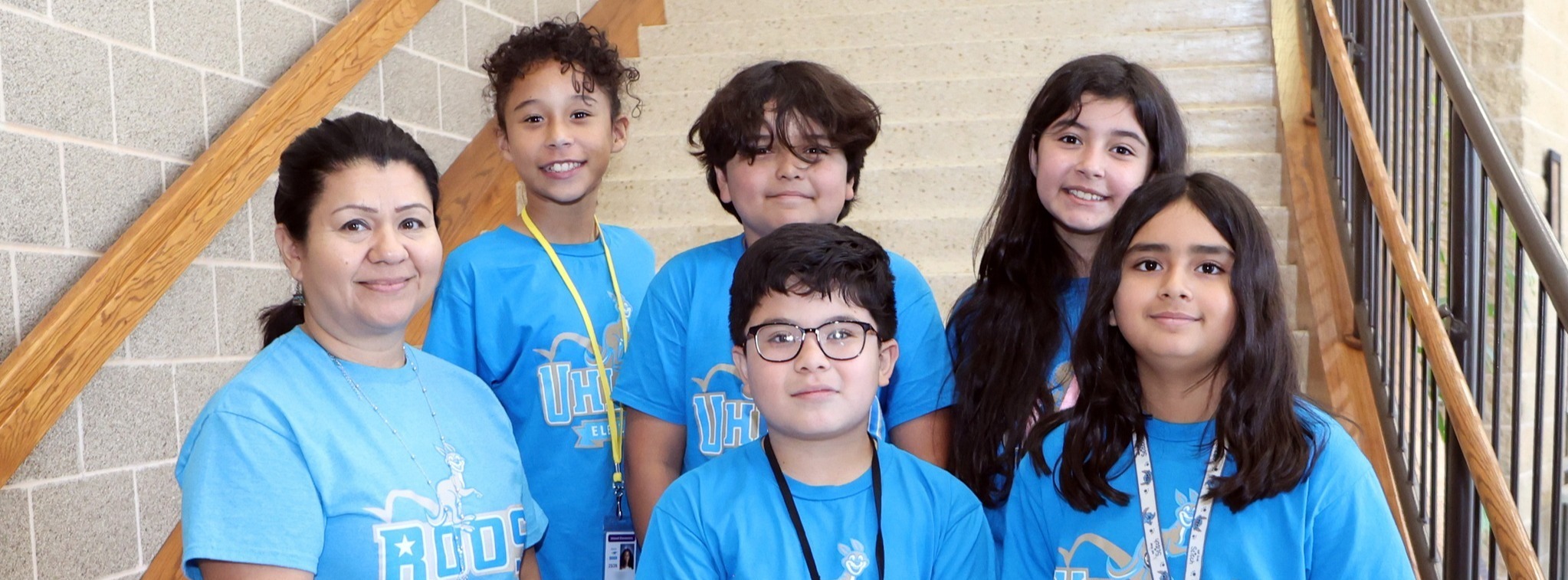 A group of five smiling students and a teacher wearing light blue matching shirts while posing for a group photo on a set of stairs.