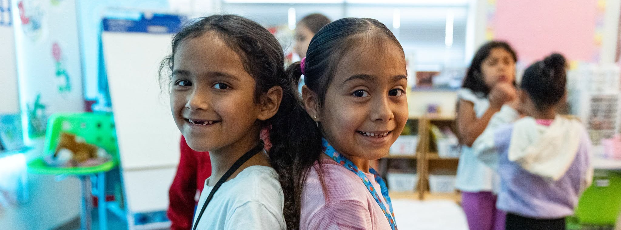 Two young girls standing back to back while looking at the camera and smiling.