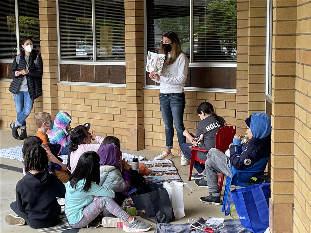A person is reading to a group of children outdoors, gathered on the ground with some seated in chairs.