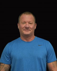 A man with short hair and tattoos, wearing a blue athletic shirt, smiles at the camera against a black background.