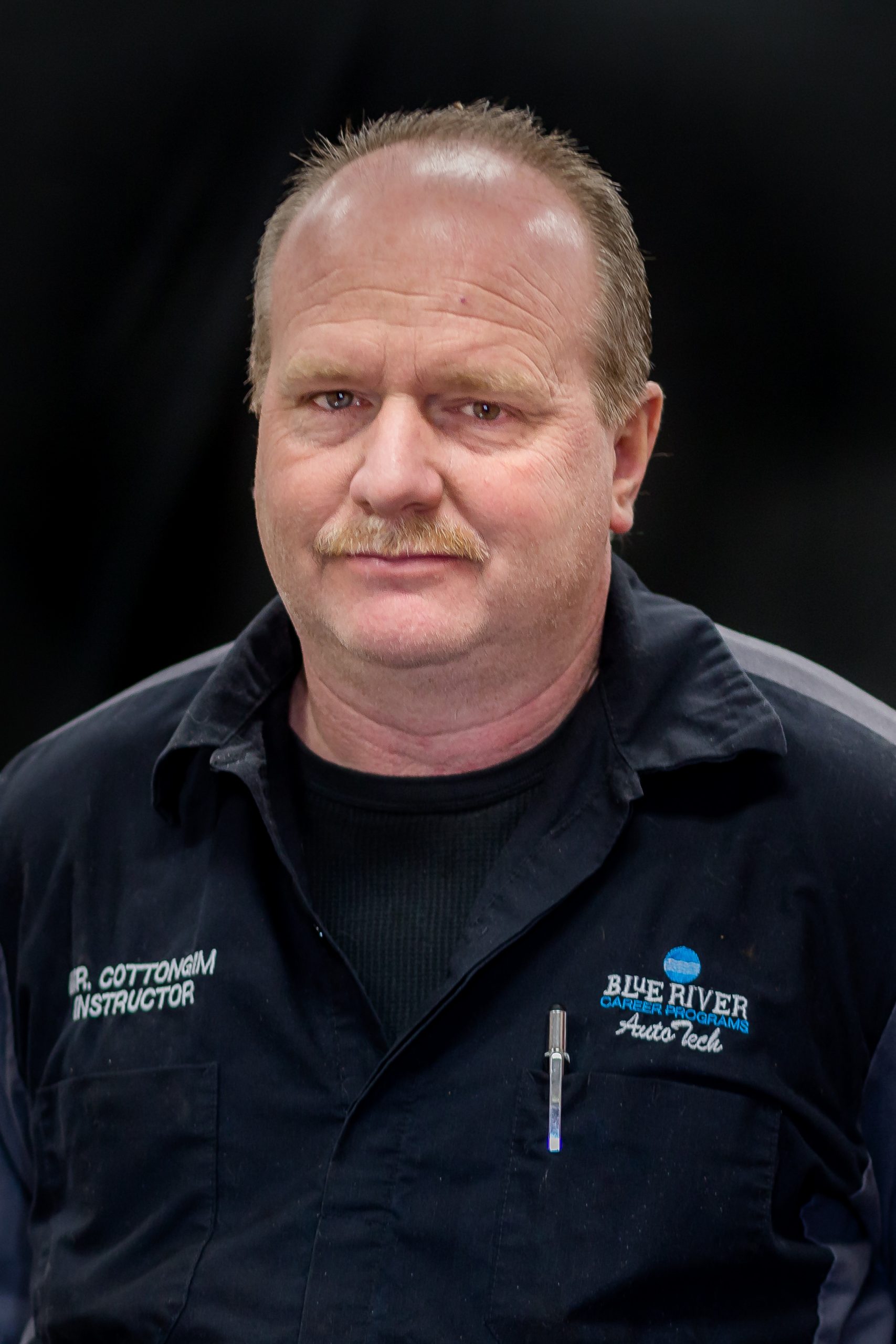 A middle-aged man with short hair and a mustache, wearing a dark blue work shirt with the name 'G. Gutmann' and a logo for 'Blue River Auto Tech.'