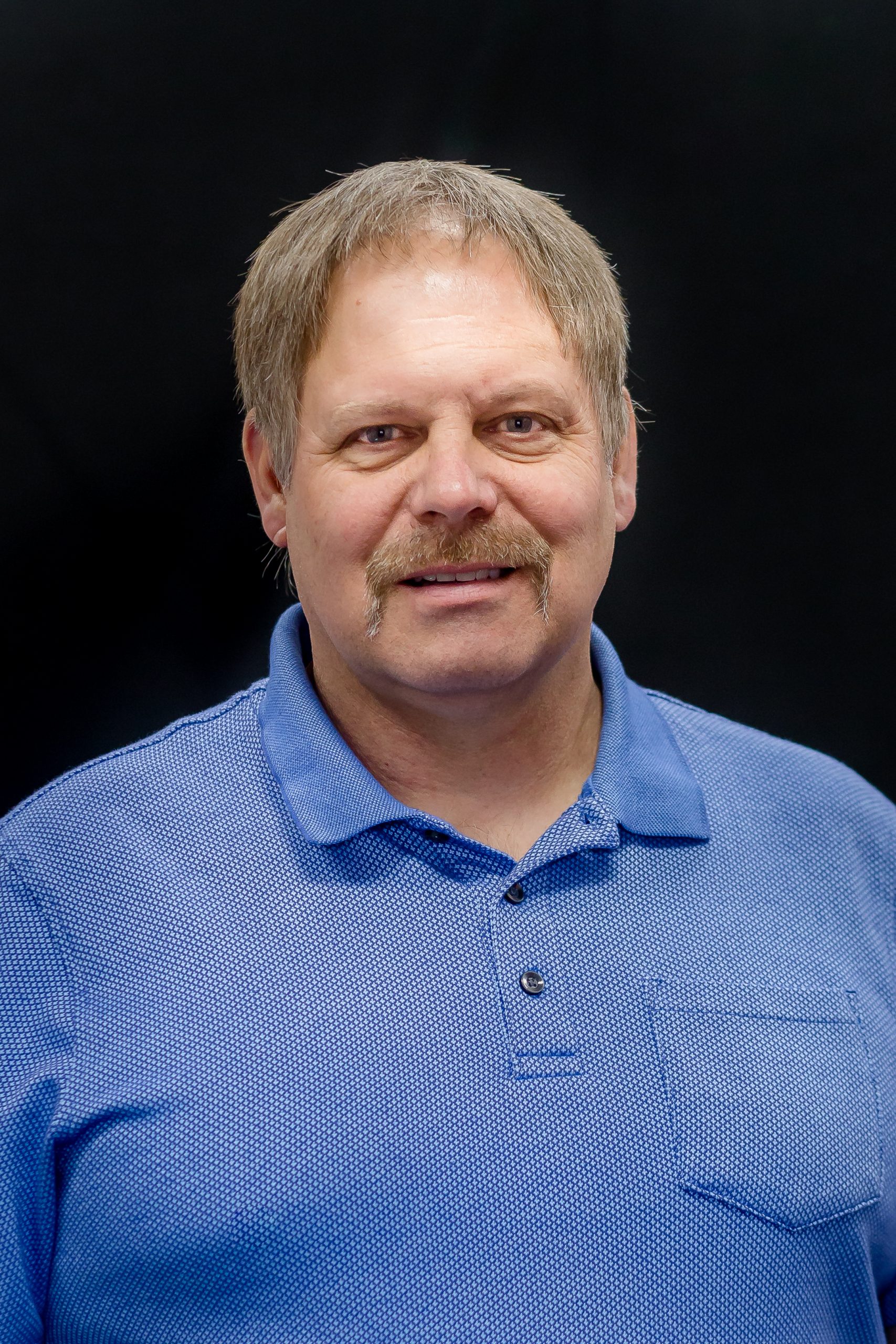 A man with short, light brown hair and a mustache is smiling while wearing a blue polo shirt against a dark background.