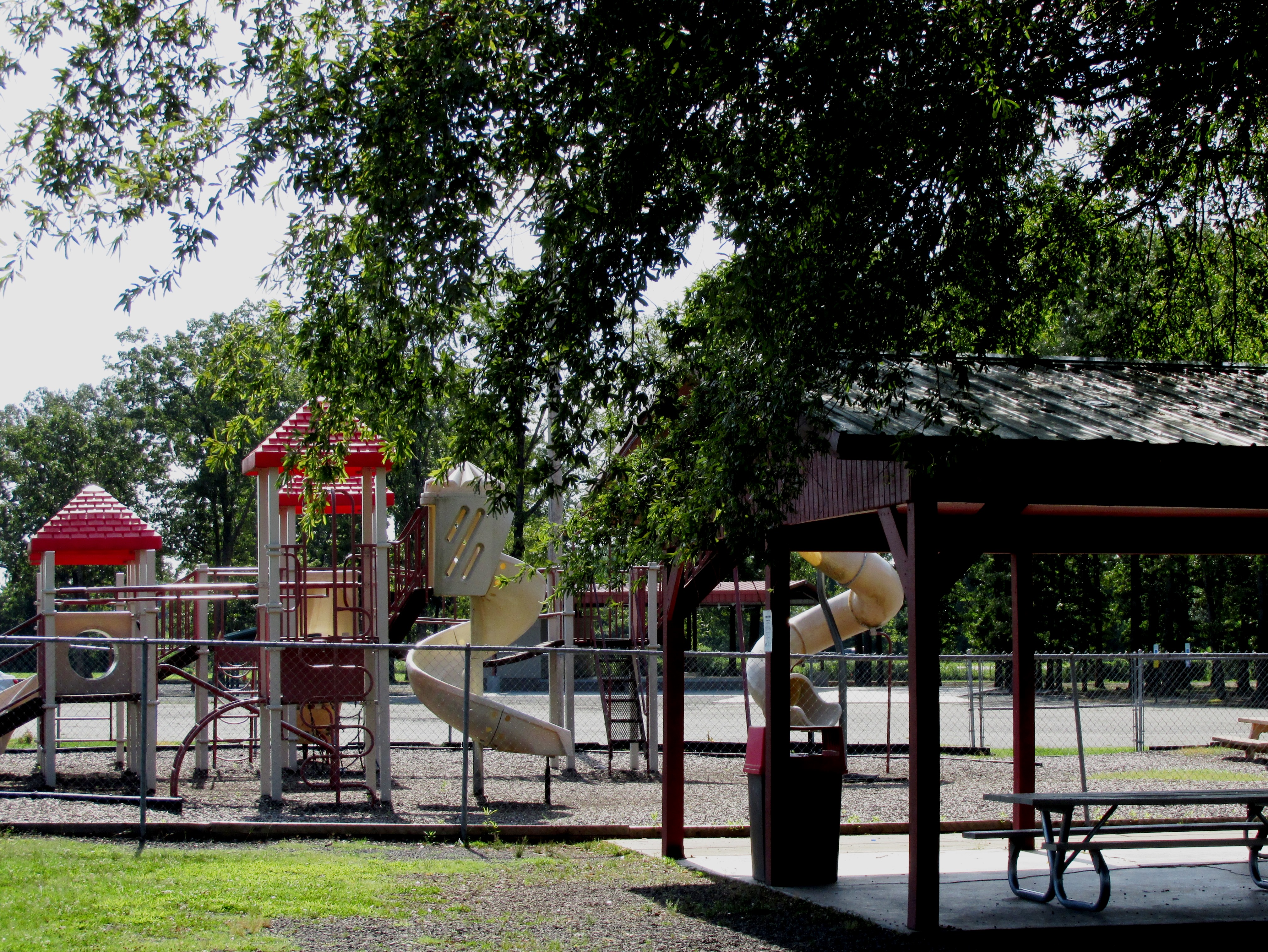 A playground featuring colorful slides and climbing structures, with a shaded picnic area nearby, surrounded by trees.