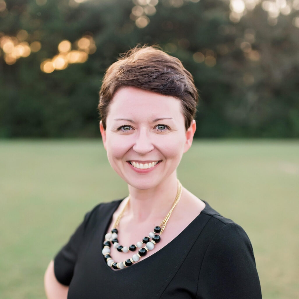 A woman with short hair smiles confidently while standing outdoors in a grassy area, wearing a black dress and a statement necklace.
