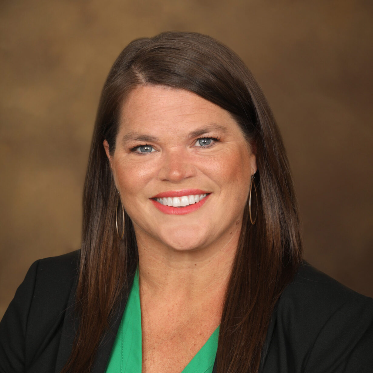 A smiling woman with long brown hair, wearing a black blazer over a green top, poses for a professional headshot against a brown background.