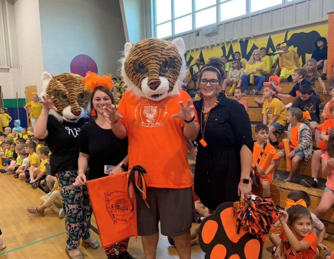 Three individuals, two wearing tiger mascot costumes and one in an orange shirt, pose together in a gym filled with children dressed in yellow and orange for a school event.