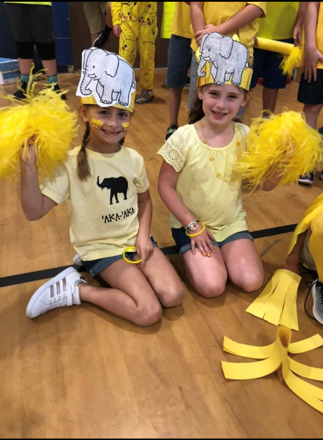 Two young girls wearing yellow shirts and elephant-themed hats are sitting on a gym floor, each holding yellow pom-poms, surrounded by other children in a festive atmosphere.