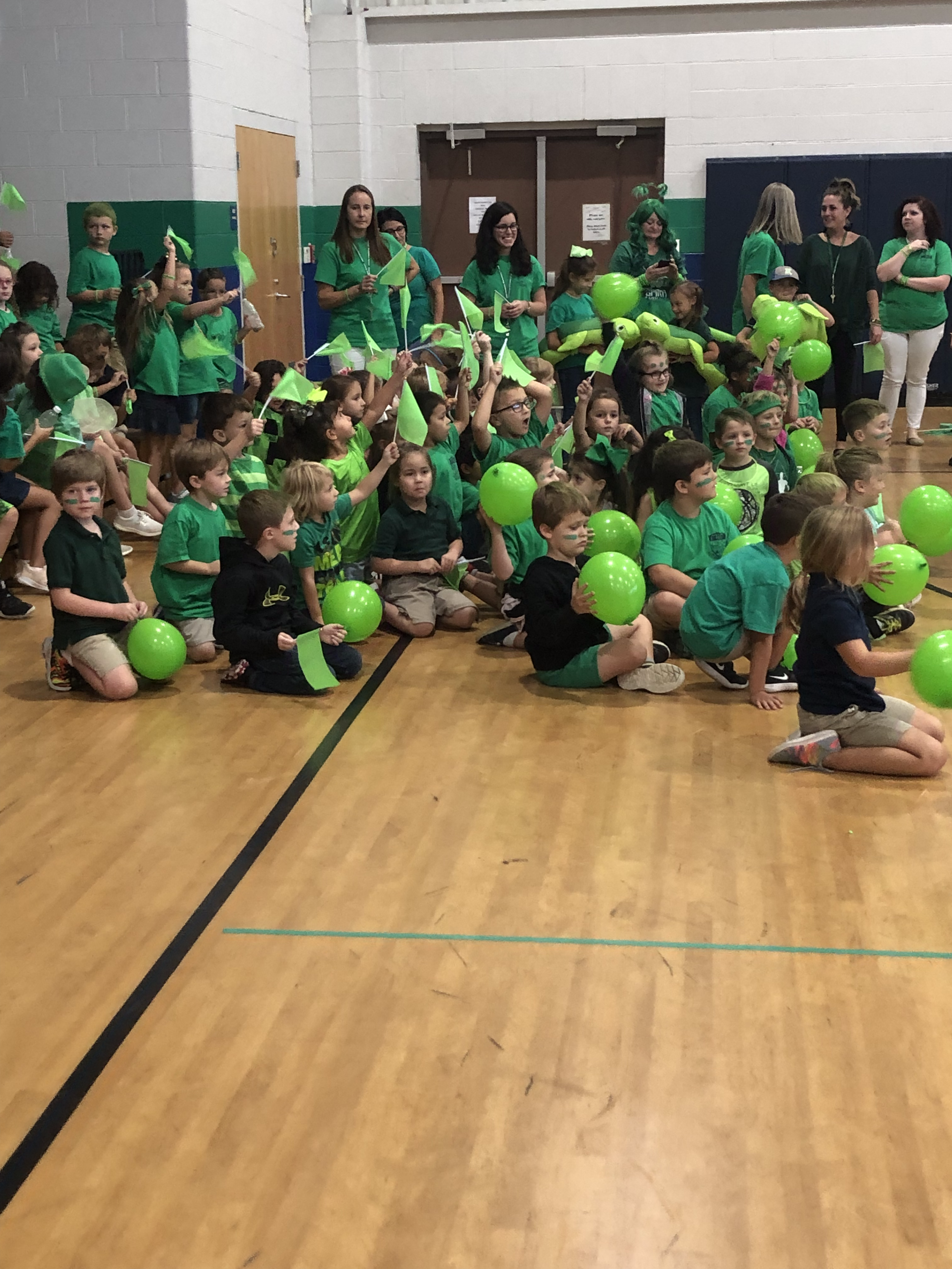A large group of children and adults, mostly wearing green shirts, are gathered in a gymnasium, holding green balloons and cheering enthusiastically.