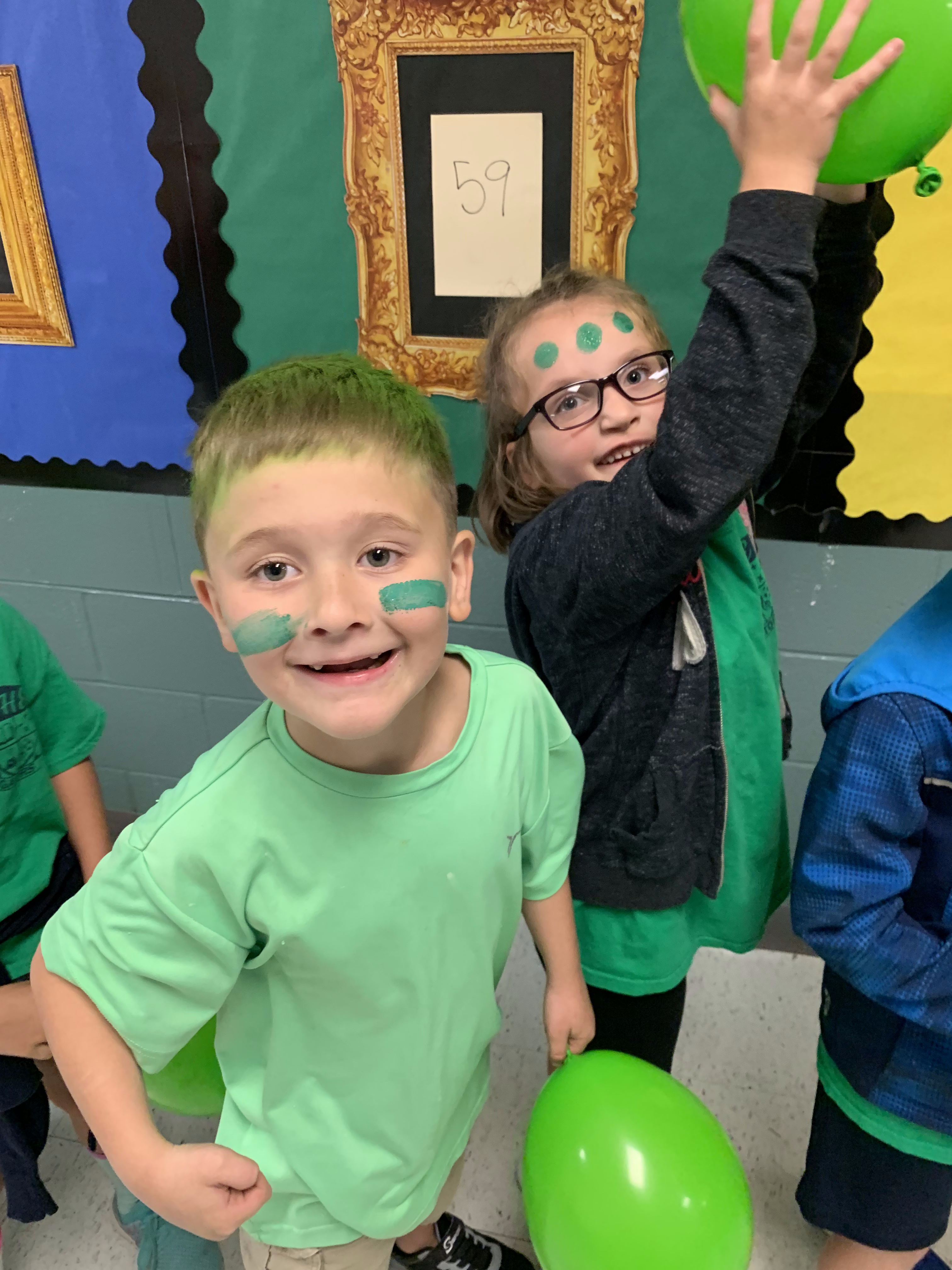 Two children dressed in green, one with face paint and holding a green balloon, and the other wearing glasses and raising a hand, are smiling in a colorful classroom setting.