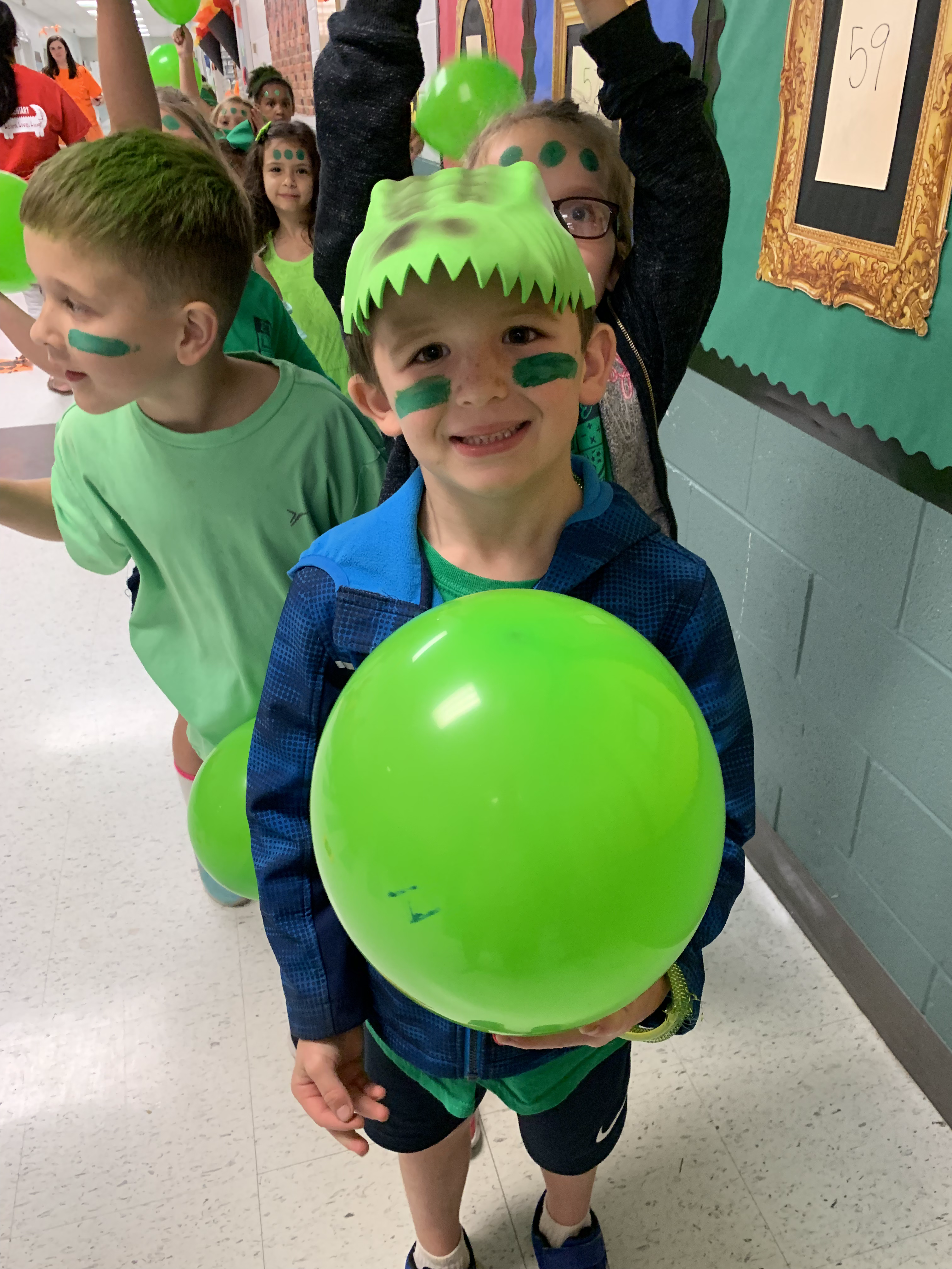 A young boy wearing a green dinosaur hat and face paint, holding a large green balloon, smiles while standing in a hallway filled with other children dressed in green.