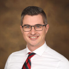 A smiling man with short, styled hair and glasses, wearing a white dress shirt and a red and black striped tie, poses against a blurred brown background.