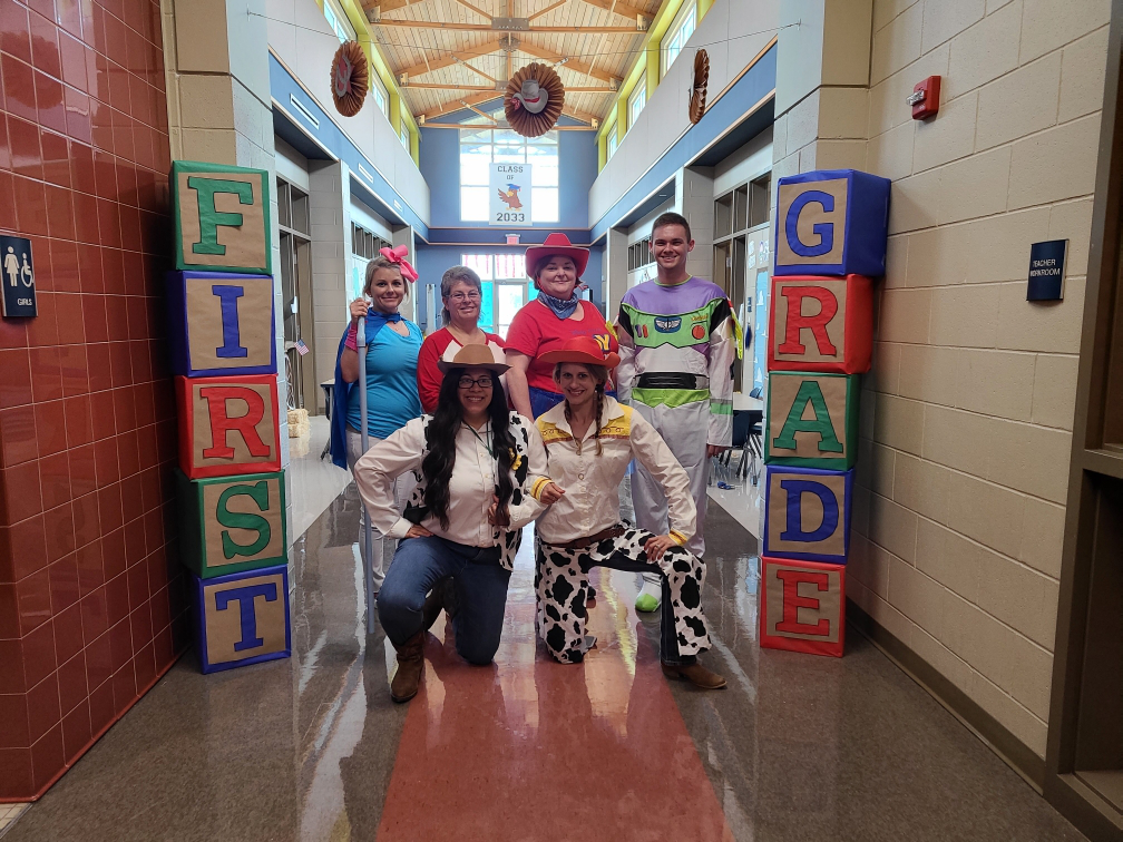A group of six individuals dressed in colorful costumes stands in a school hallway, framed by large blocks spelling 'FIRST GRADE'.