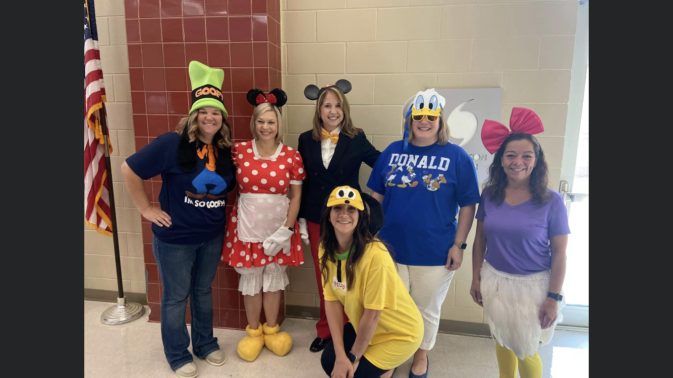 A group of six women dressed in colorful costumes inspired by Disney characters, including Minnie Mouse and Donald Duck, posing together in a school setting with an American flag in the background.