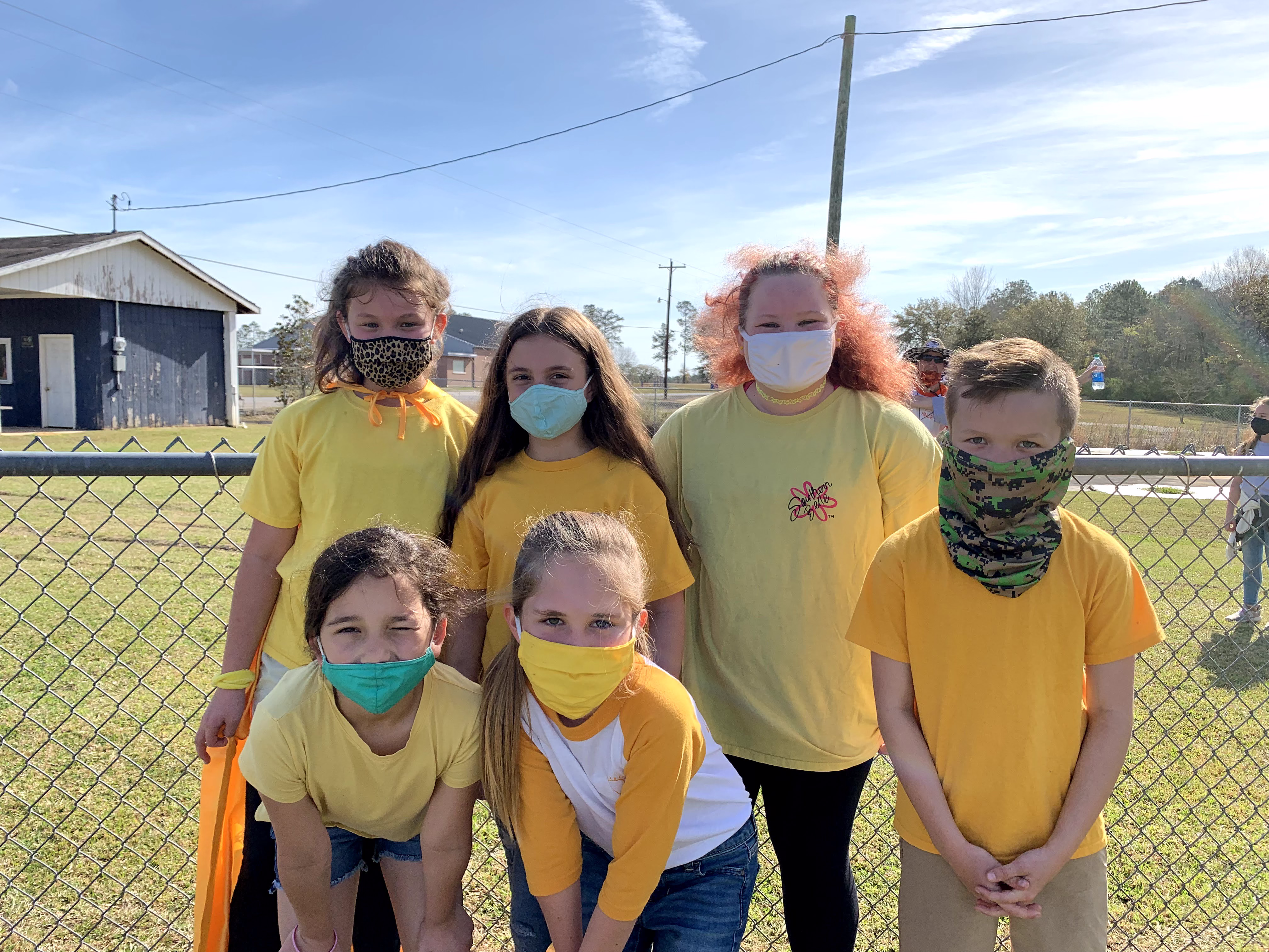 A group of six children, wearing yellow shirts and various face masks, pose together outdoors in front of a fence on a sunny day.