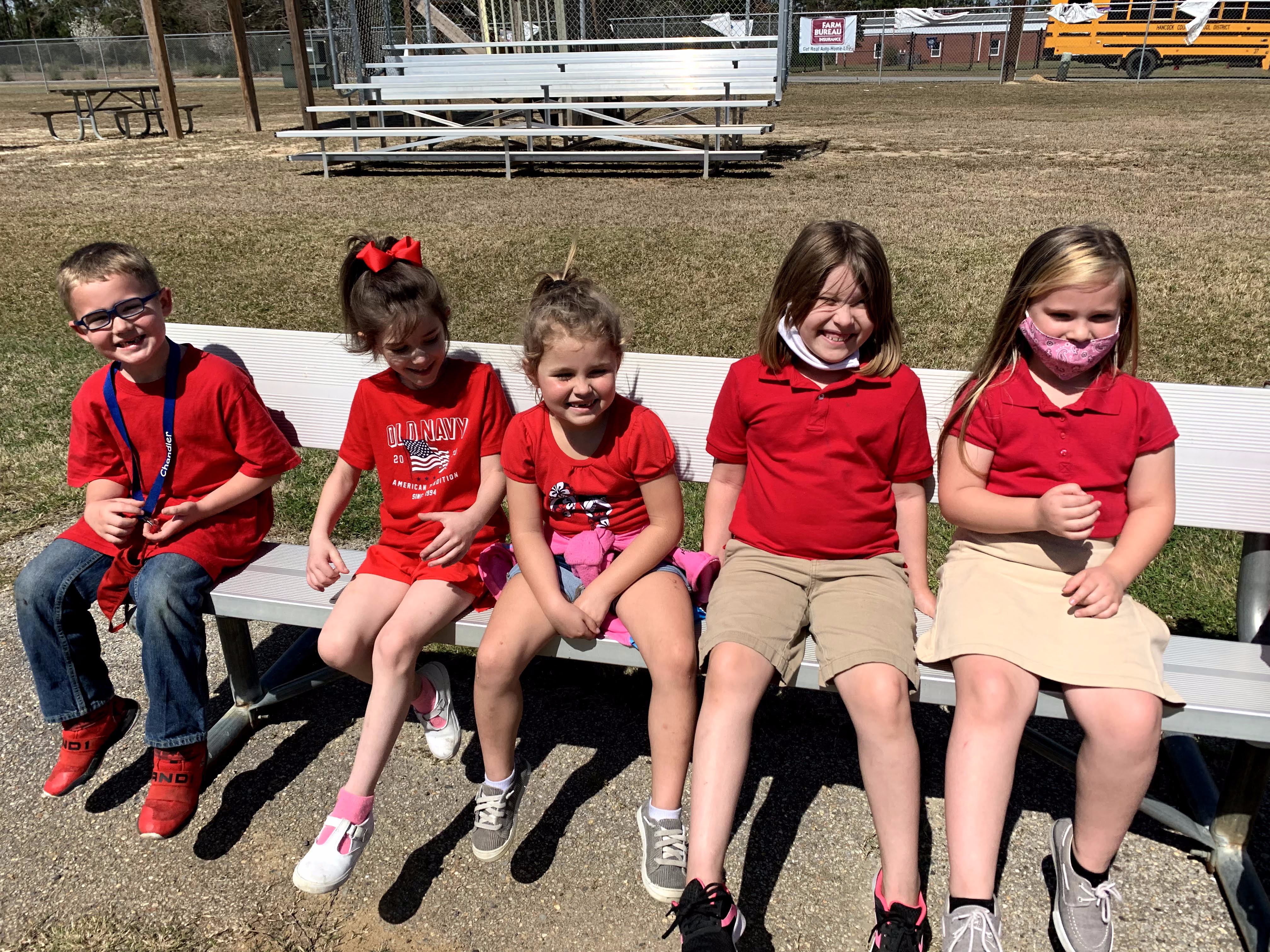 Five children wearing red shirts sit together on a bench in a grassy area, smiling and enjoying a sunny day.