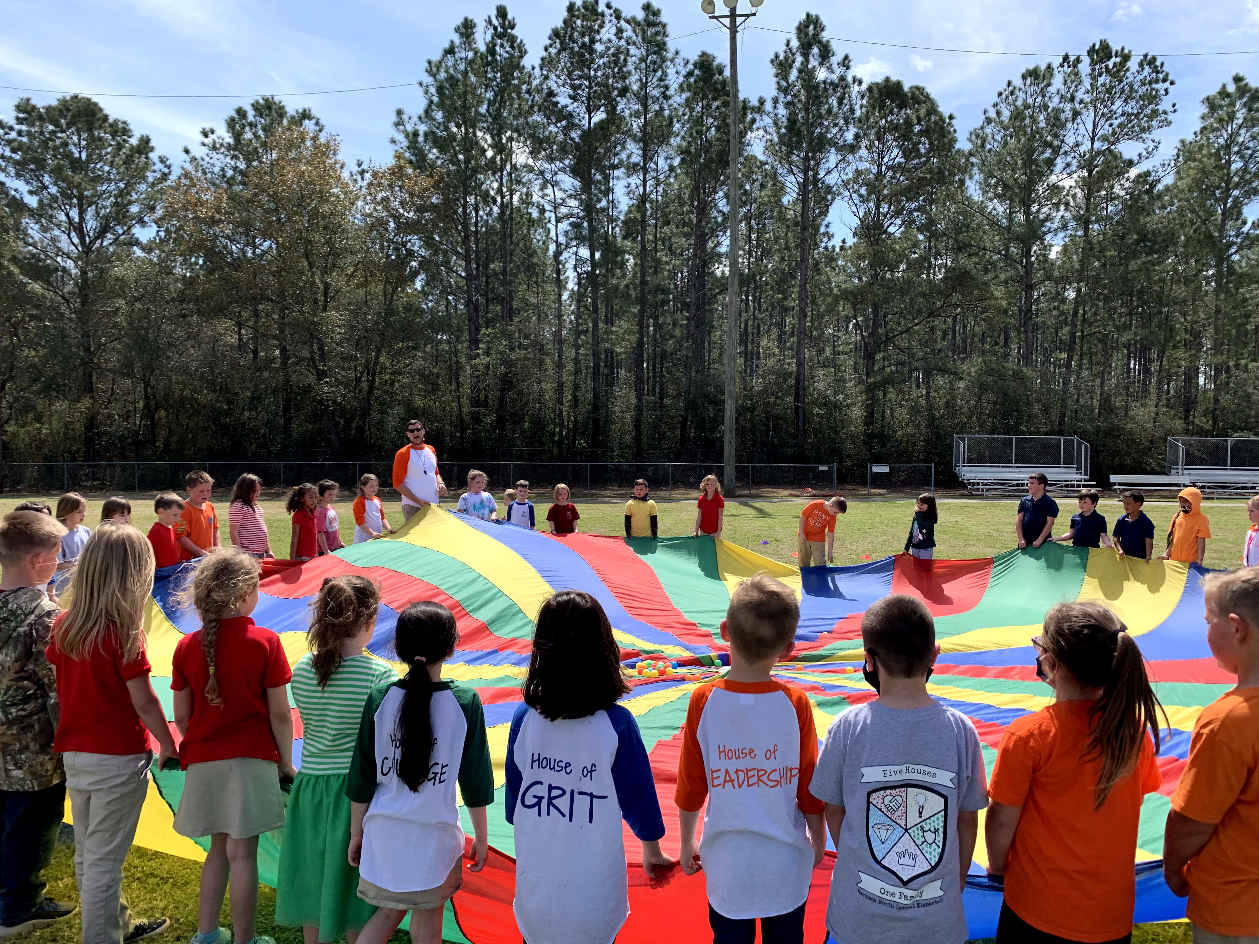 A group of children in colorful shirts are gathered around a large, vibrant parachute on a grassy field, with trees in the background, as they participate in a playful activity led by an adult.