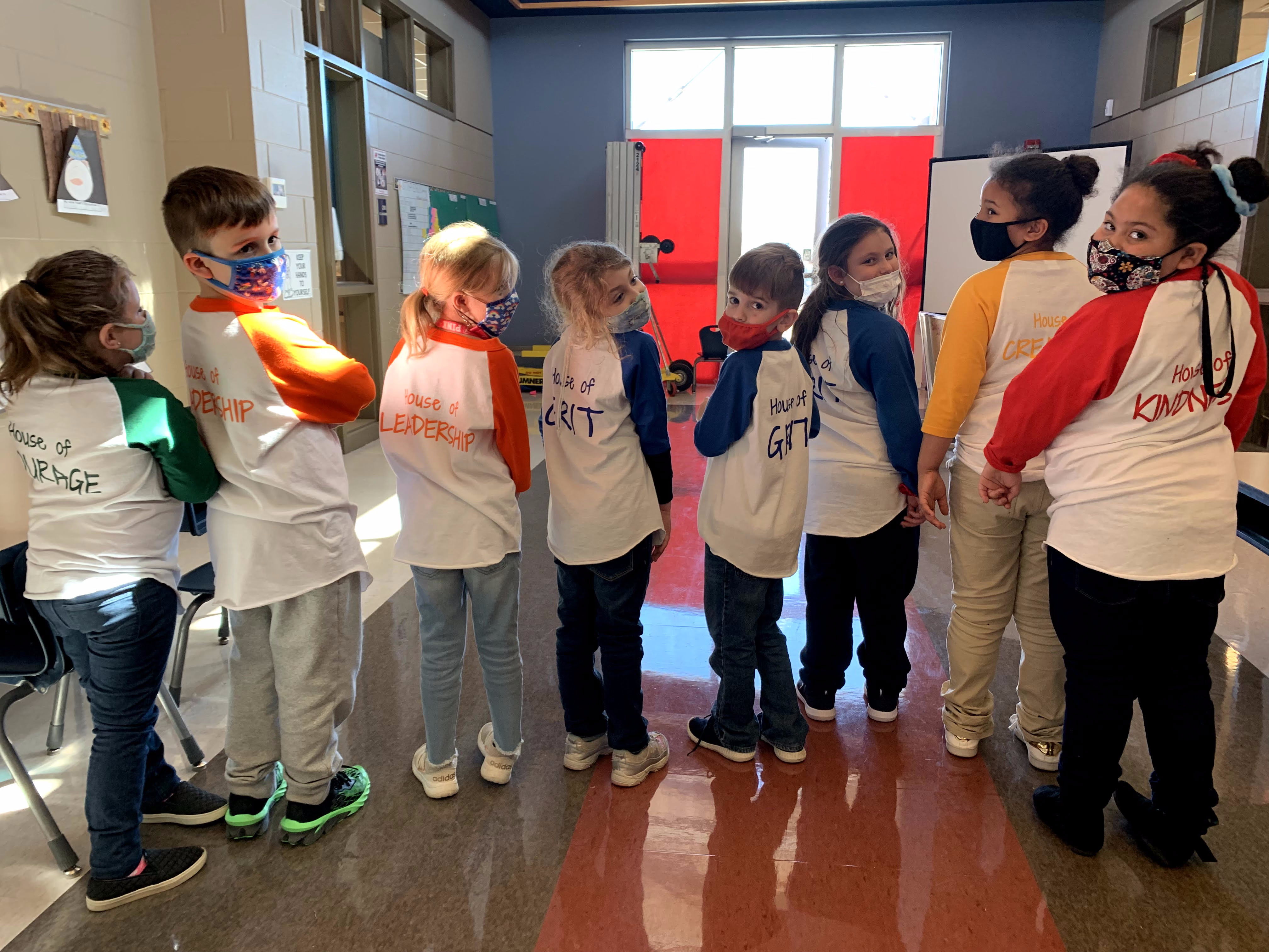 A group of children wearing matching white t-shirts with colorful sleeves stand in a line, facing away from the camera, in a brightly lit hallway.