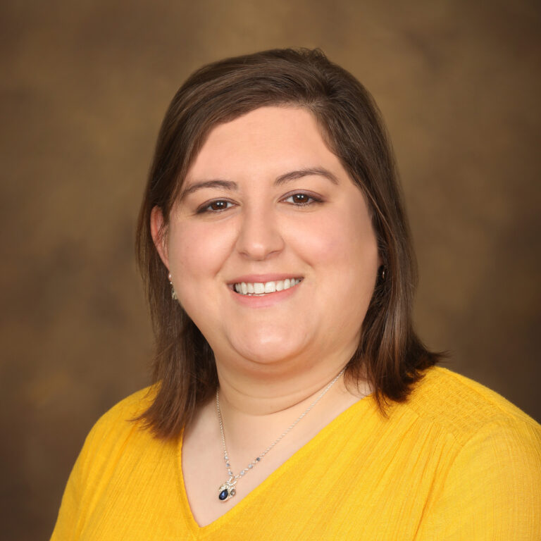A smiling man with short, styled hair and glasses, wearing a white dress shirt and a red and black striped tie, poses against a blurred brown background.