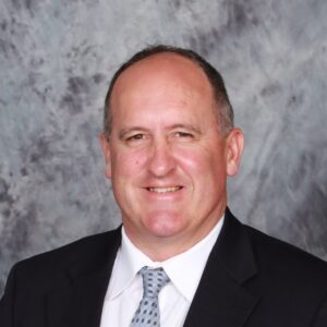 A man in a suit and tie smiles at the camera, set against a textured gray background.