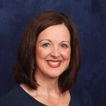 A woman with shoulder-length brown hair smiles warmly against a dark blue background, wearing a navy blue top.
