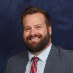 A smiling man with a beard, wearing a gray suit and a red-striped tie, poses against a blue background.