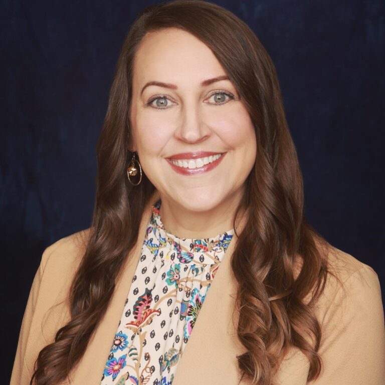 A woman with long, wavy brown hair smiles warmly at the camera, wearing a beige blazer over a floral blouse, set against a dark background.