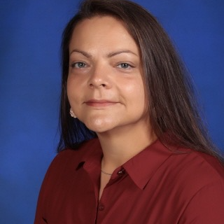A woman with long dark hair and a serious expression is wearing a maroon blouse against a blue background.