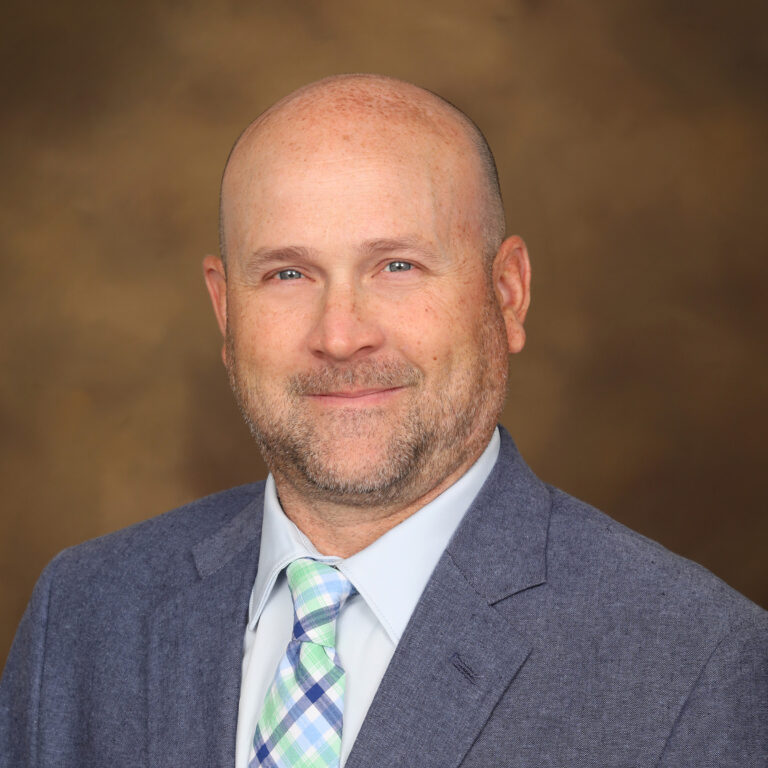 A man with a bald head and a short beard, wearing a blue blazer over a light blue shirt and a green and blue plaid tie, smiles against a blurred brown background.